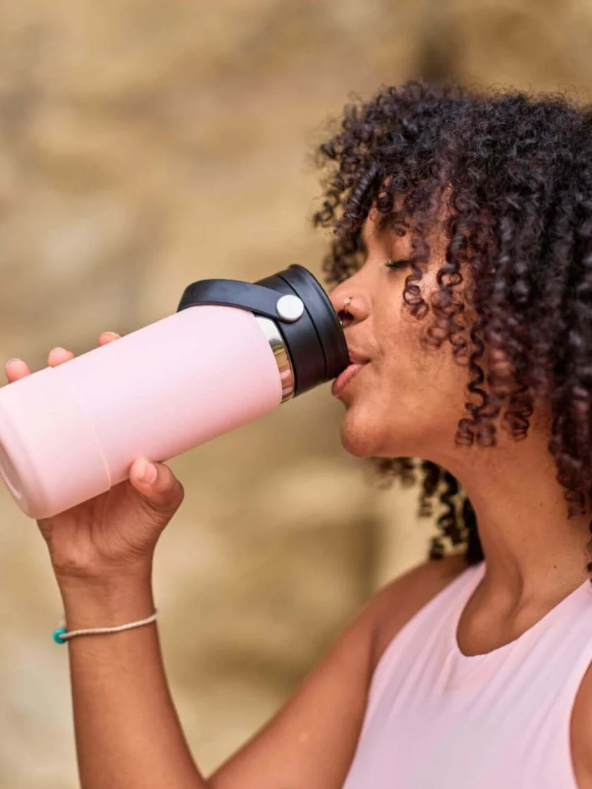Una mujer con el pelo rizado y una camiseta de tirantes rosa bebe de una botella de agua Hydro Flask de 20 oz con tapa Flex Sip Lid.