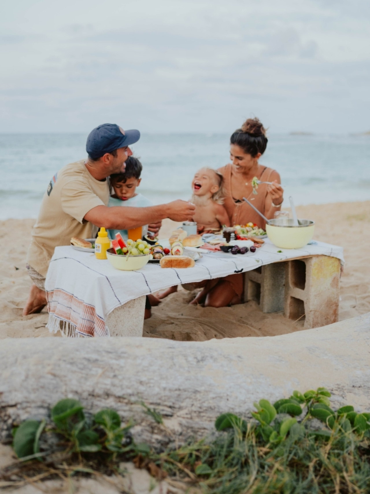 A happy family of four enjoying a beach picnic with veggie skewers and food served in Hydro Flask Outdoor Kitchen bowls and plates on a wooden table covered with a white blanket, with the waves and sand in the background. The parents, in their early 30s w