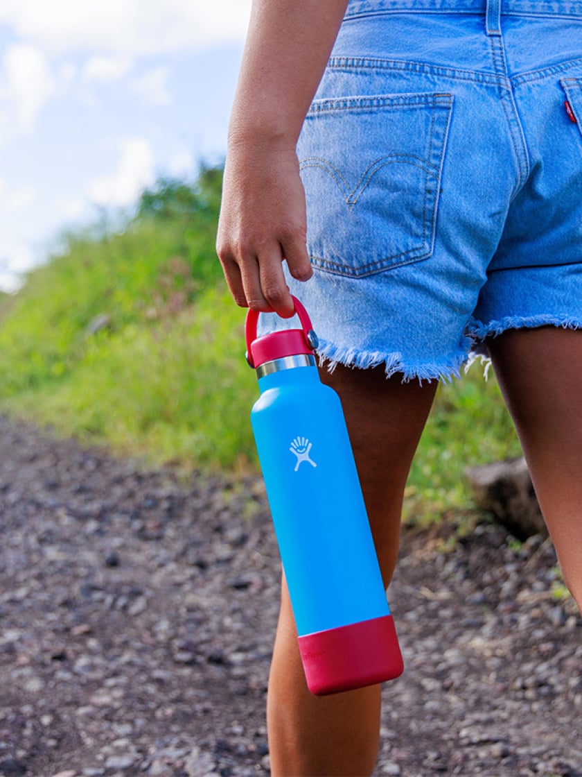 A person wearing jean shorts walks along a tropical trail, holding a Hydro Flask bottle with a custom cap  and bottle boot.