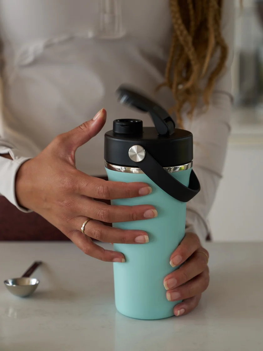 A woman wearing athletic wear is opening a light blue Insulated Shaker Bottle to prep her workout drink.