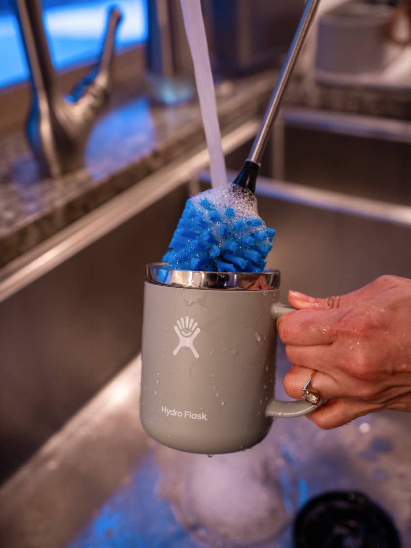 Hydro Flask mug held under running water in sink while being scrubbed with a bottle brush