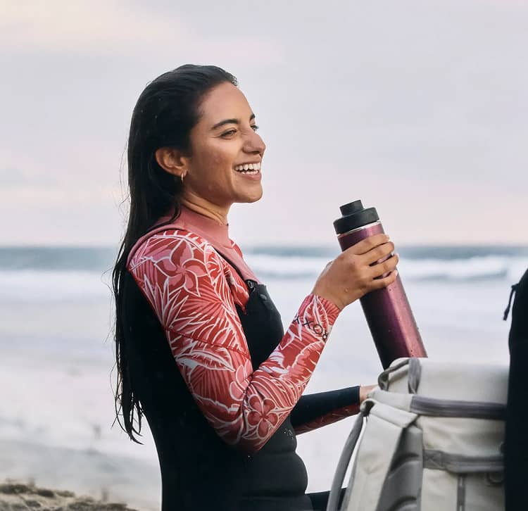 Girl in wet suit smiling and drinking out of a lightweight hydro flask on the beach with a surfboard